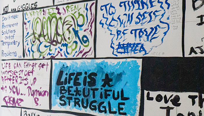 Photo of a wall at seperated into brick-style rectangles. Each one has personal painted messages on it. The most prominent reads 